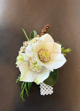 Load image into Gallery viewer, A small floral boutonniere featuring a large pale cream hellebore flower with green leaves, tiny white filler blossoms, and a pearl bead cluster, arranged on a dark brown background.

